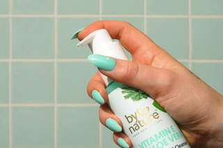 Hand holding a 'by nature' vitamin e cleanser against a tiled wall background