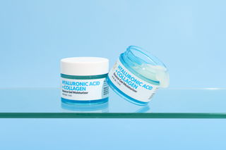 Two cosmetic jars of Hyaluronic Acid + Collagen moisturizer on a clear glass shelf against a light blue background. One jar is upright and closed with a white lid, and the other is tipped over to show the pale blue gel inside.