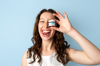 A woman with long brown hair smiles while holding a small cosmetic jar over one eye against a light blue background.