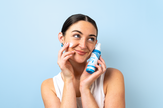 A woman with dark hair pulled back applies a white cream to her cheek while holding a blue and white spray bottle against a solid light blue background.