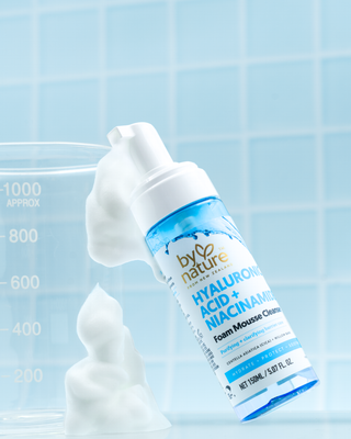 A bottle of by nature Hyaluronic Acid + Niacinamide Foam Mousse Cleanser shown with white foam against a light blue tiled background and a glass beaker.