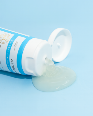 A squeeze tube of tooth gel lies on its side with the cap open, dispensing a translucent pale green gel onto a smooth light blue background. The tube is white with blue text and a small gold circular logo.