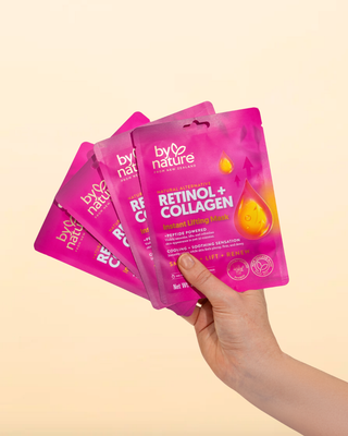 Hand holding pink 'by nature' retinol + collagen instant lifting mask packages against a beige background